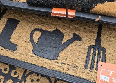 Detail kokosové předložky s vyvýšeným gumovým motivem holínky, zahradní konvičky a hrábí, s oranžovou etiketou BEST FOR BOOTS