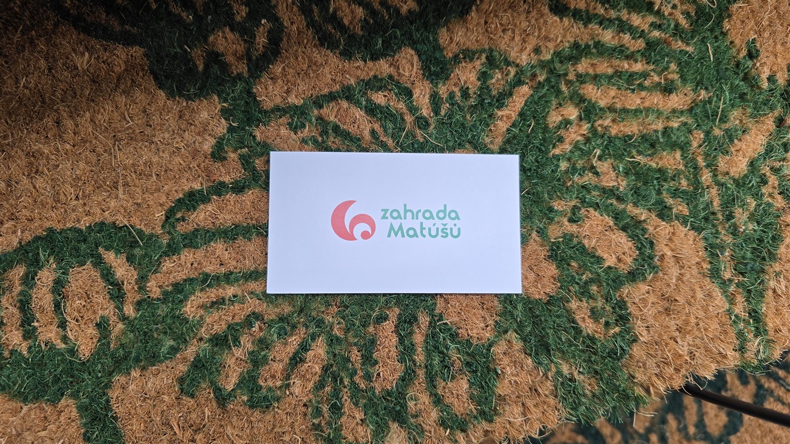 Kokosová předložka se vzorem velkých listů v zeleném a přírodním odstínu vlákna, na ní vizitka Zahrada Matúšů