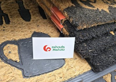 Stoh tmavě probarvených kokosových předložek vedle světlé rohožky s motivem konvičky a hrábí, s oranžovými etiketami